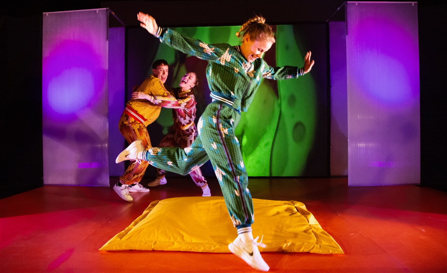 Bild från föreställningen Pirret (för 10-12 år). En skådespelare hoppar i förgrunden och i bakgrunden skrattar och kramas två skådespelare. Scenografin är färgglad.
