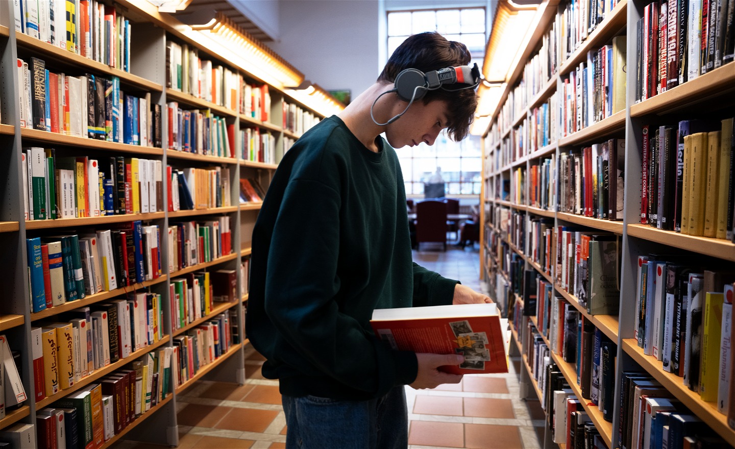 En person med hörlurar står i ett bibliotek och läser i en bok