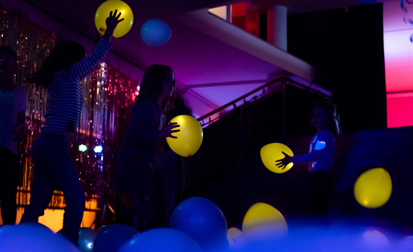 Barn dansar med självlysande ballonger. I bakgrunden syns ett glitterdraperi och ljus i olika färger.  