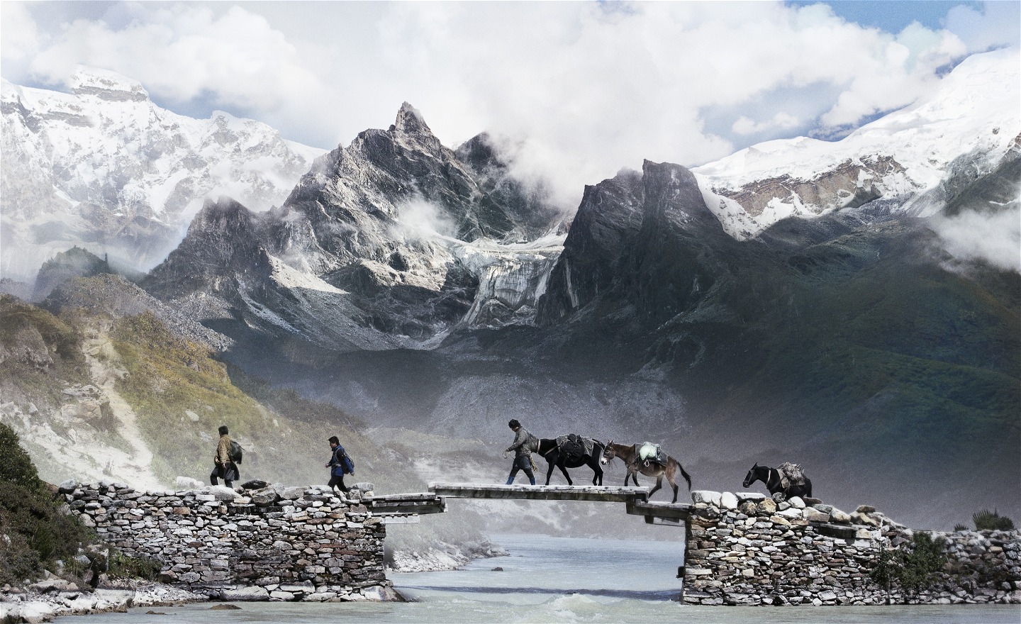 Tre människor med åsnor går över en stenbro vid älven i ett bergsmassiv i Bhutan.