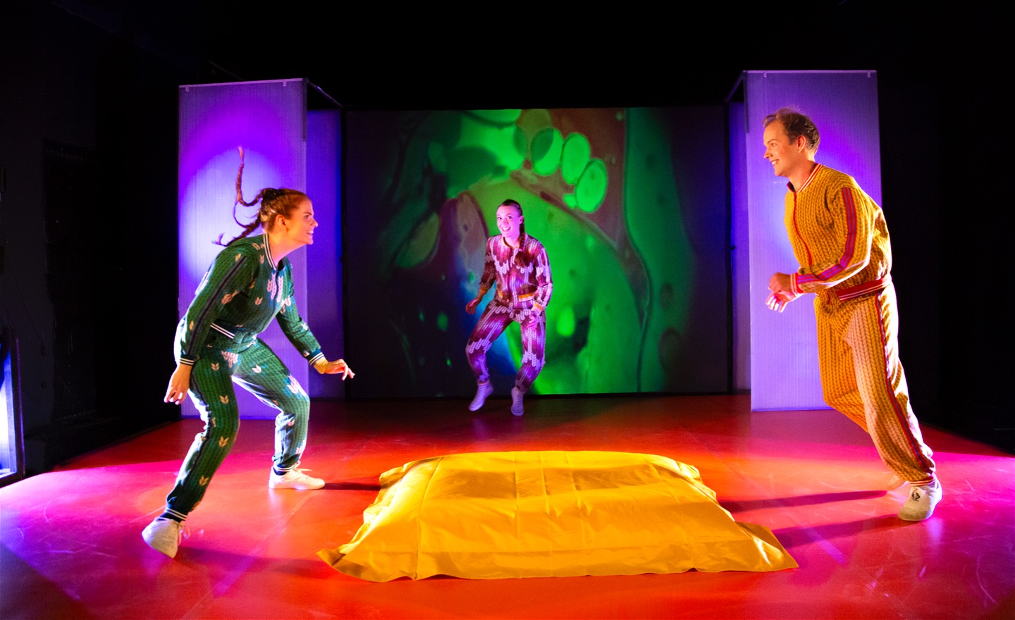 Bild från föreställningen Pirret. De tre skådespelarna jagar varandra runt en ul kudde. Scenografin är färgglad.