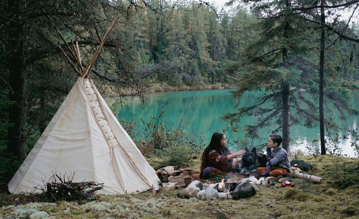 En flicka, en pojke och en hund sitter vid ett tält och en lägereld i vildmarken.