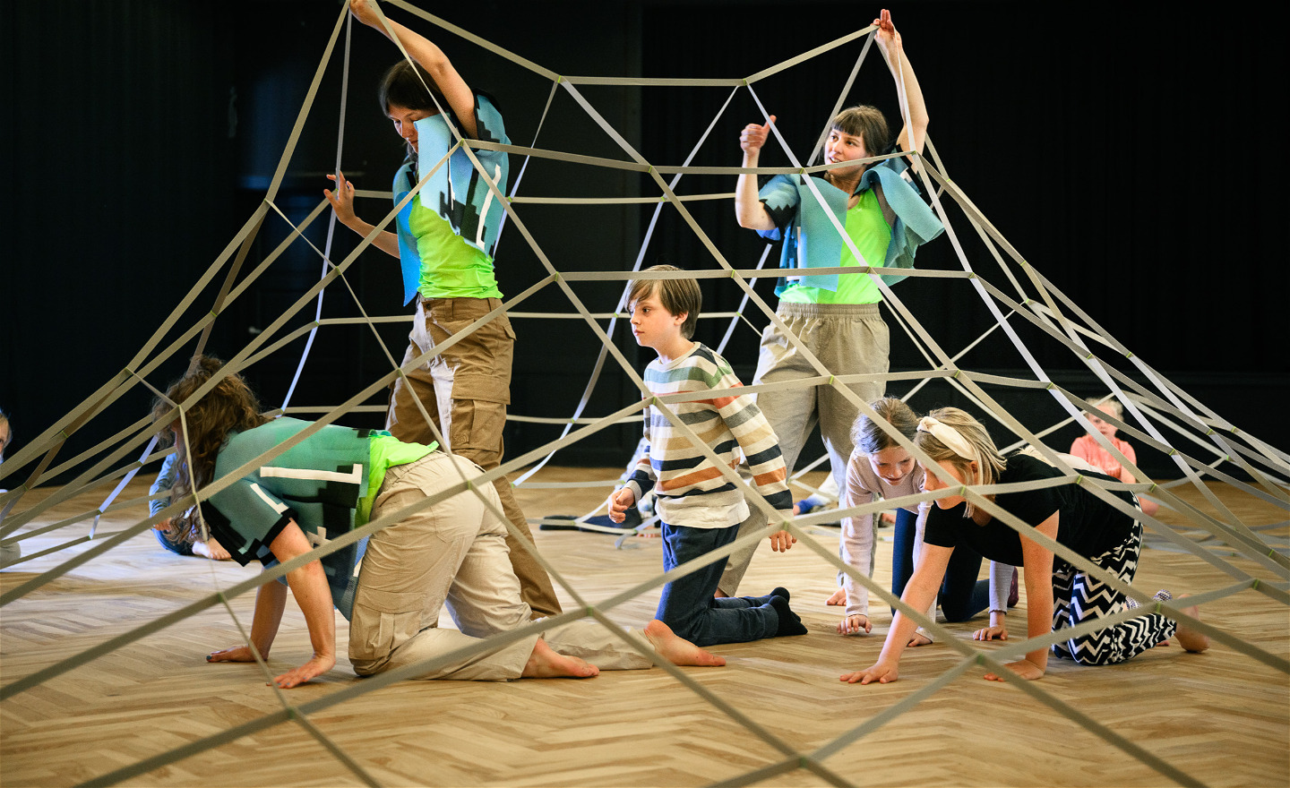 Två dansare håller upp ett stort elastiskt nät som flera barn och en dansare kryper under. 