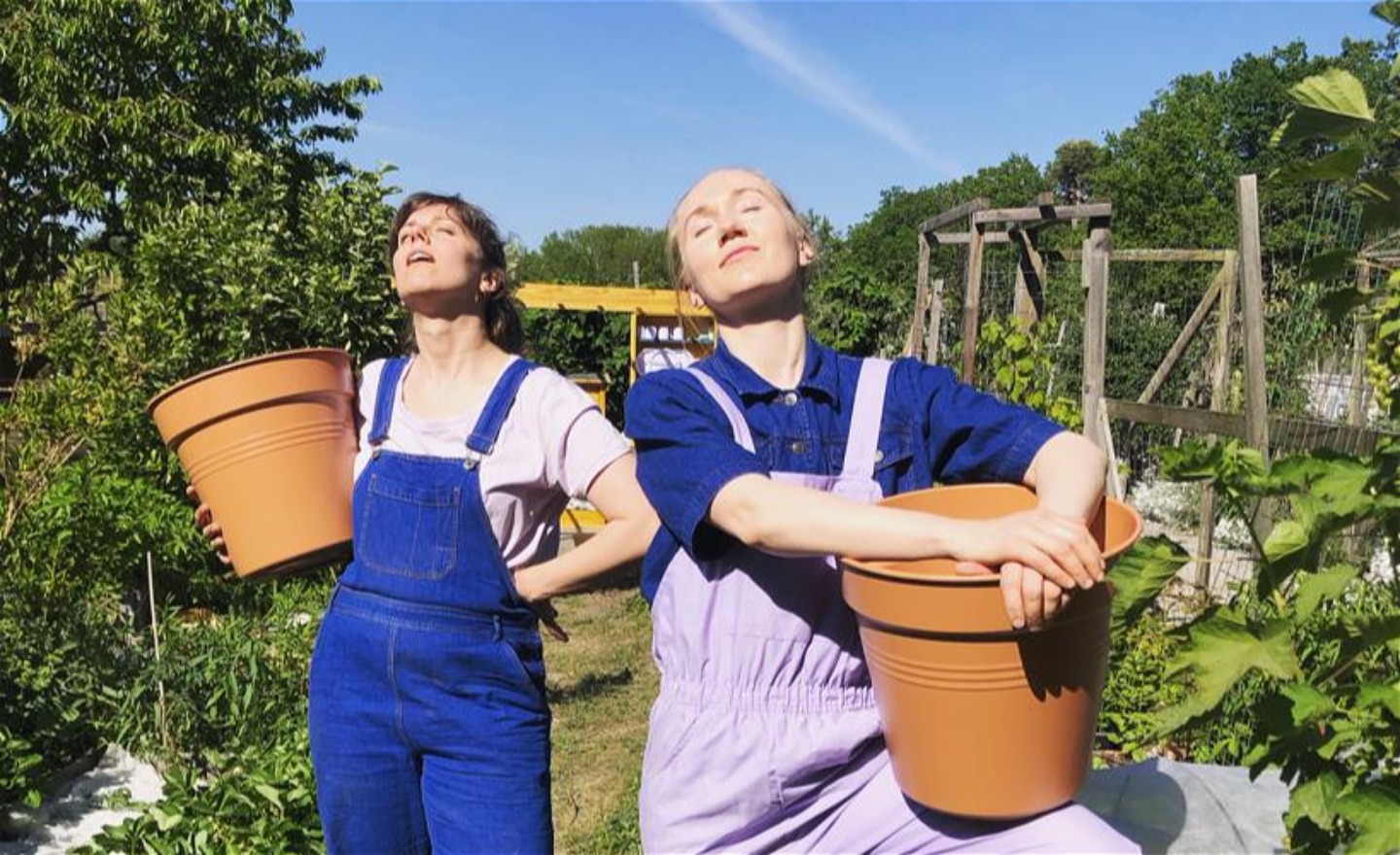 Två trädgårdsmästare solar ståendes i en trädgård, de håller i varsin stor kruka