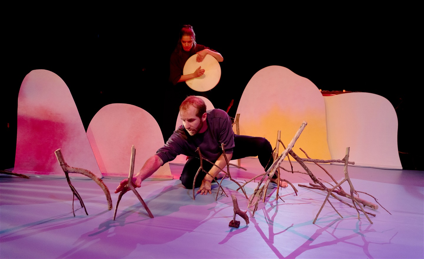 En man placerar grenar på en scen, en kvinna står bakom och slår på en trumma
