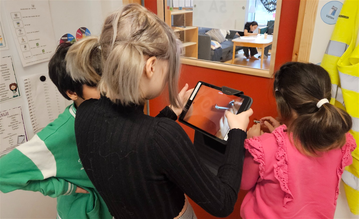 Tre barn står i en halvcirkel, barnet imitten håller en iPad och filmar när barnet till höger rycker i dörrhandtaget