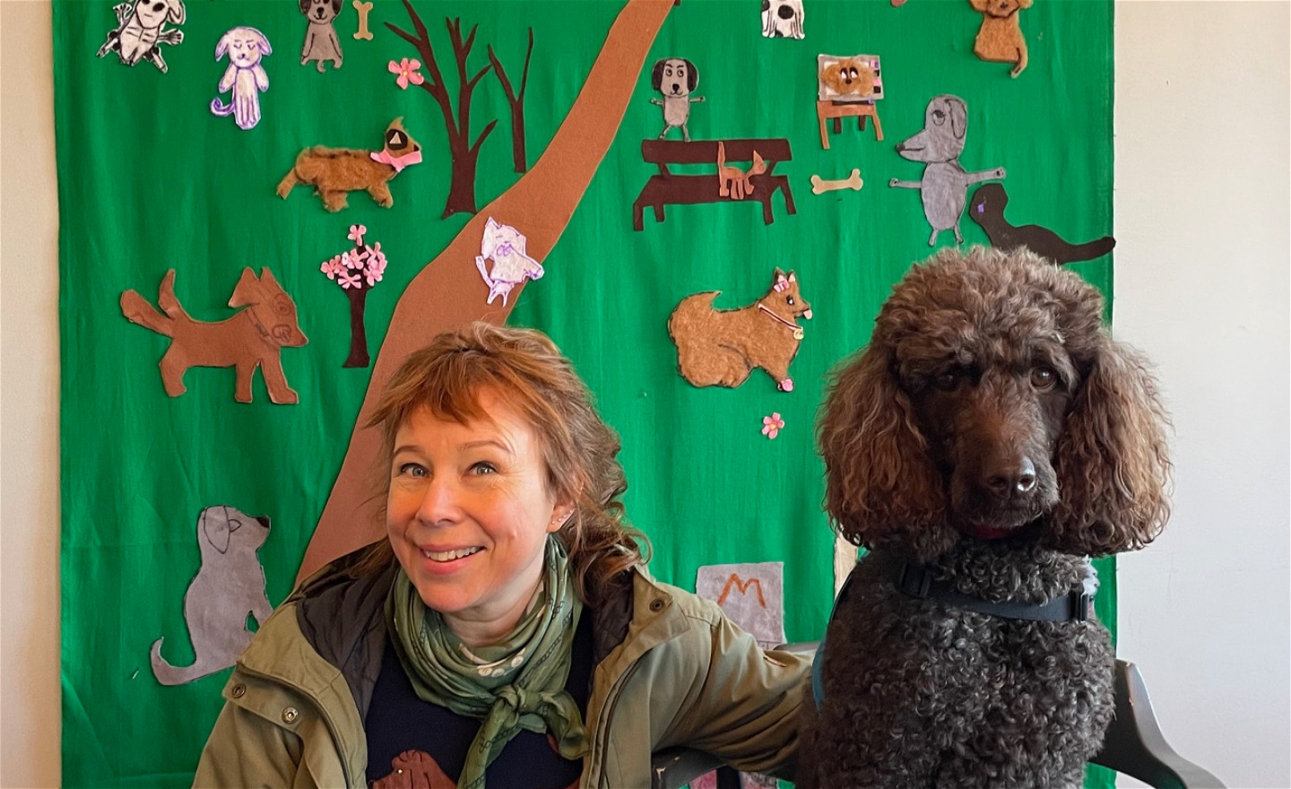 Konstnären Catti Brandelius och hunden Kokkos poserar glatt framför en tygbonad prydd av hundar