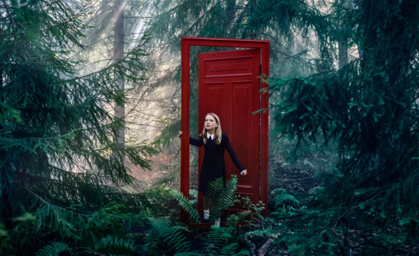 Ett barn öppnar en röd dörr mitt i en granskog och tittar sig omkring. Flickan har en mörk klänning på sig och har långt blont hår.