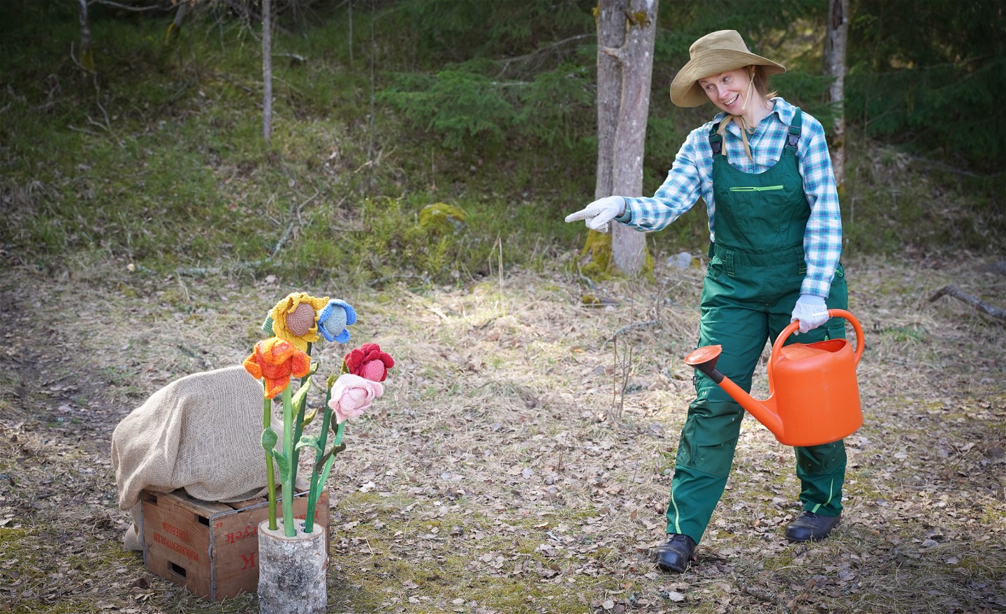 Färgglada virkade blommor och en trädgårdmästare som pekar på blommorna med ena handen. Den andra håller i en orange vattenkanna