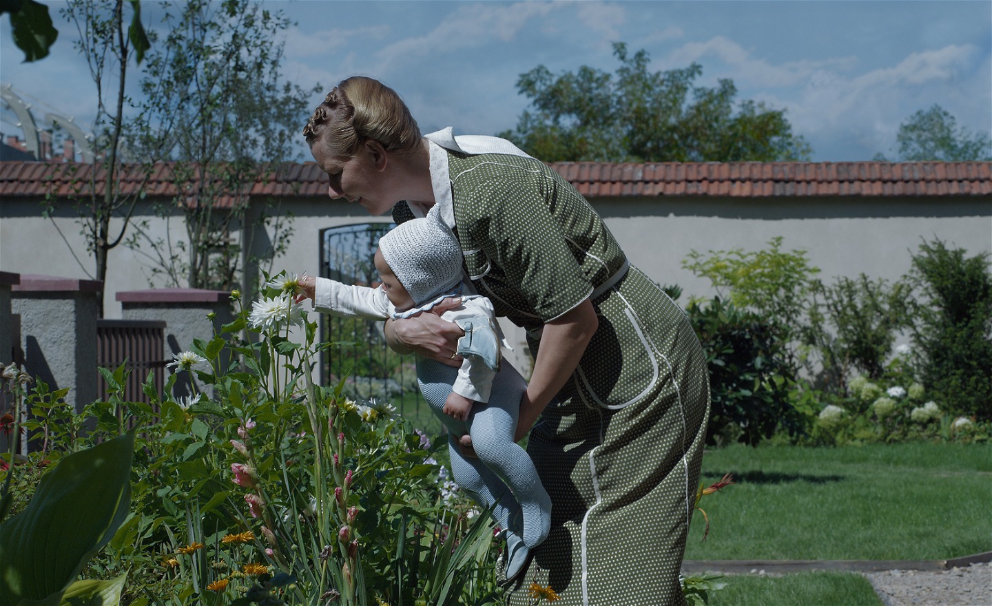 Kvinna i grön klänning står i en trädgård. Hon håller i en bebis som känner på en blomma som växer i rabatten. Bakom dem syns muren till ett koncentrationsläger.