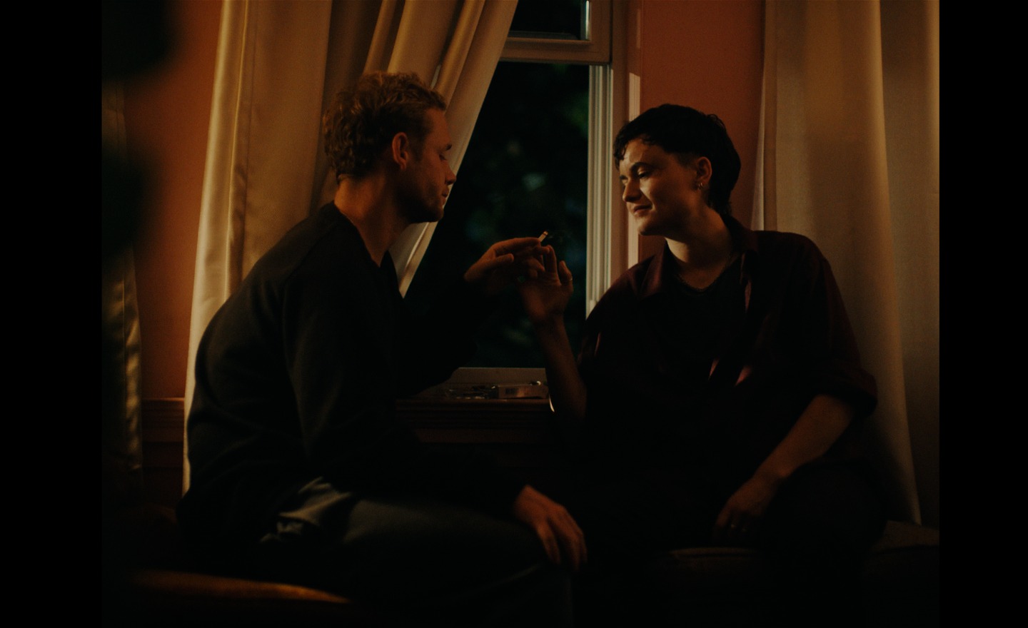 Man och kvinna sitter framför fönstret i ett dunkelt rum med varmt ljus. De delar på en cigarett. 