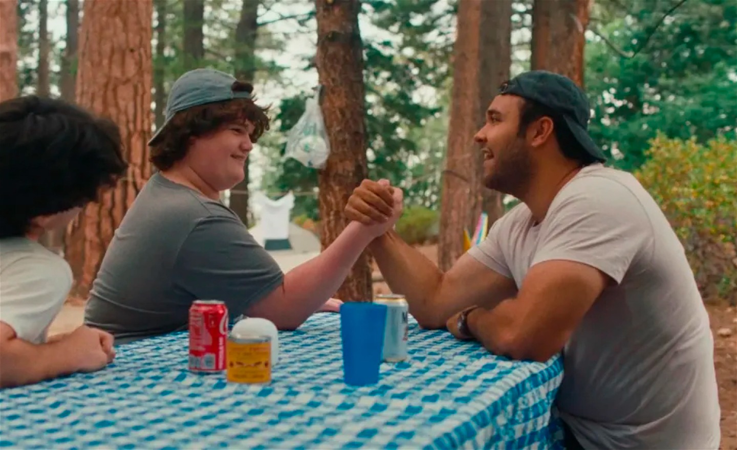 Två tonårspojkar pojkar och en man sitter vid ett picknick-bord i skogen. Ena pojken bryter arm med mannen. Båda ser glada ut.