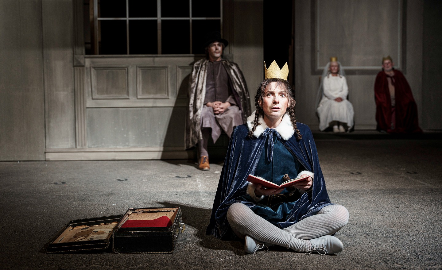 Bilden föreställer en scen i föreställningen Titta Hamlet. Hamlet sitter ner och läser boken Titta Hamlet, bakom honom sitter hans mamma, hans styvpappa och Ofelias pappa. 