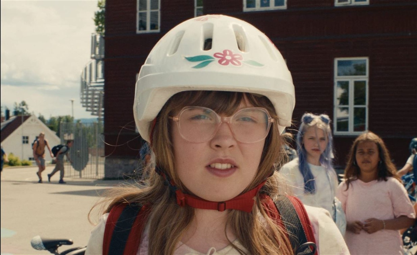 Allvarlig flicka iförd cykelhjälm, glasögon och ryggsäck står på en skolgård med andra elever i bakgrunden.