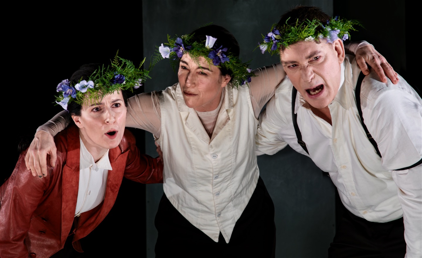 Tre personer står med kransar i håret och håller armarna om varandra och pratar högt.