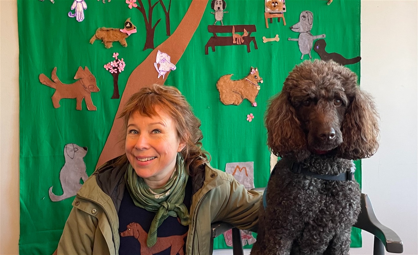 Konstnären och hundföraren Kokkos står tillsammans med den bruna storpudeln Kokkos framför en textil bild med hundar, som de har gjort tillsammans med barn.