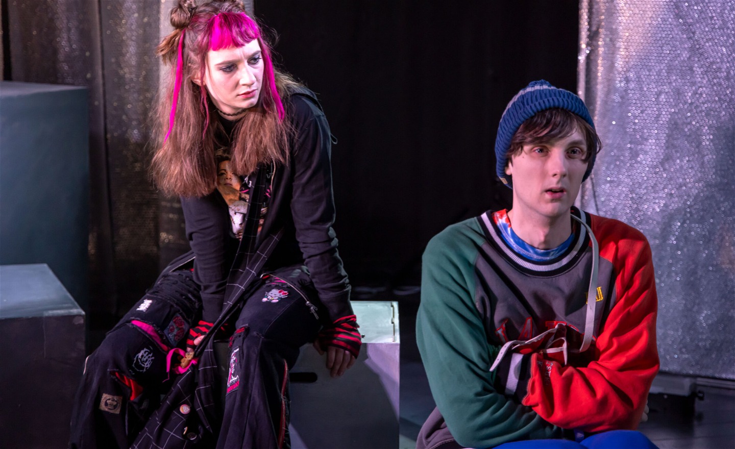 En tjej med Emokläder och rosa slingor i håret sitter på en trälåda och tittar på en kille som ser ledsen ut 