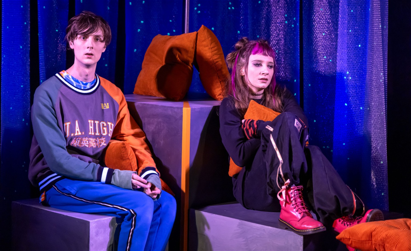 En tjej och en kille sitter på varsin trälåda och tittar rakt fram. De ser eftertänksamma och ledsna ut.  