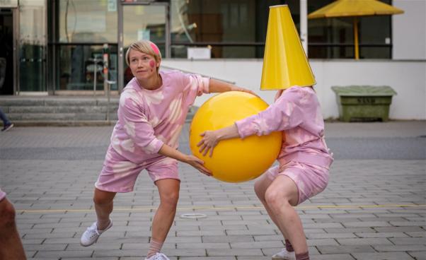Två rosaklädda dansare utomhus. De har en stor gul boll mellan sig. En har en stor, gul kon över huvudet. 