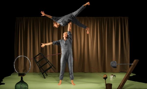 En akrobat lyfter den andra akrobaten högt upp i luften med en arm. I bakgrunden skulpturer av vardagsföremål. 